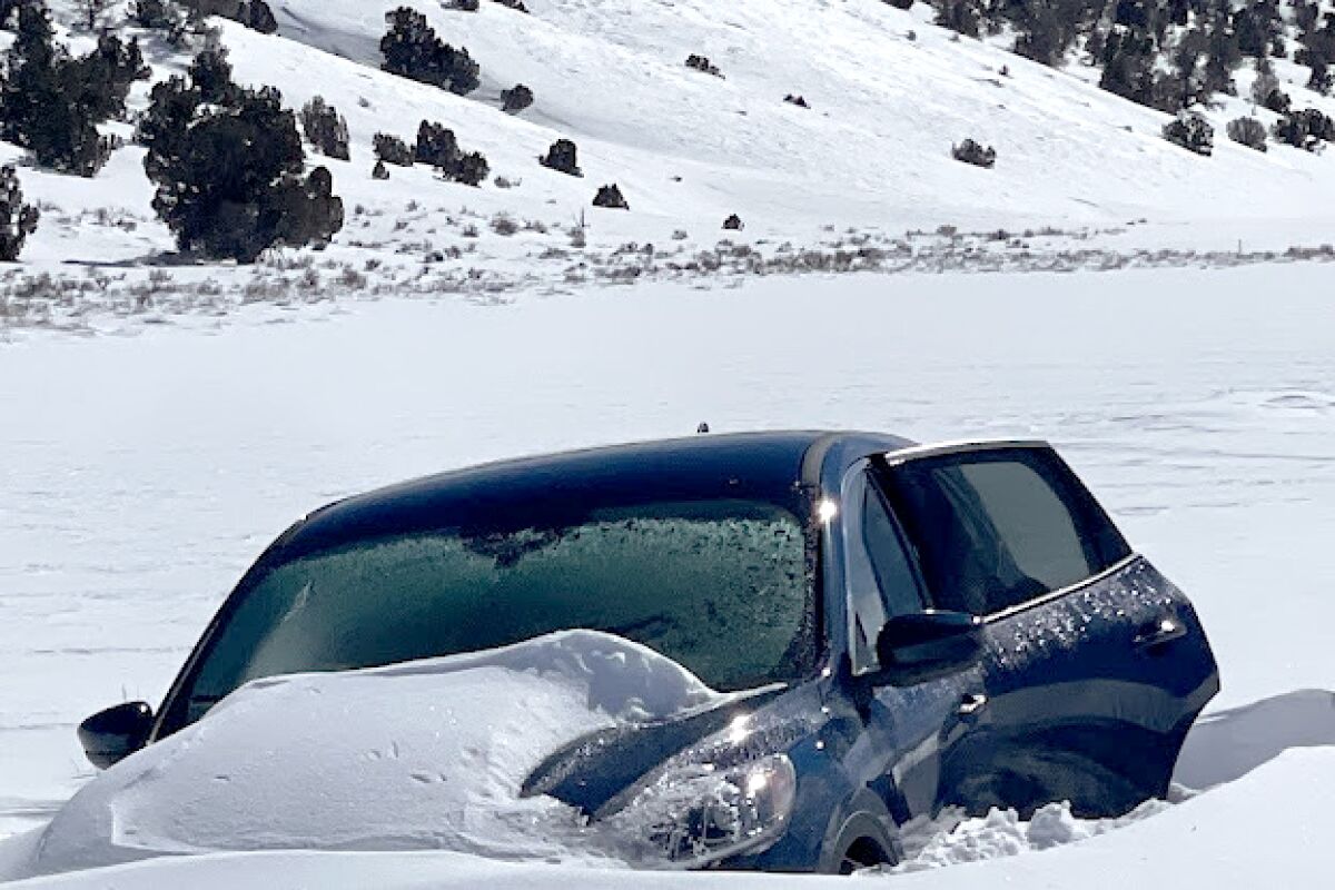 A dark-blue sedan buried in snow near a hill on horizon.
