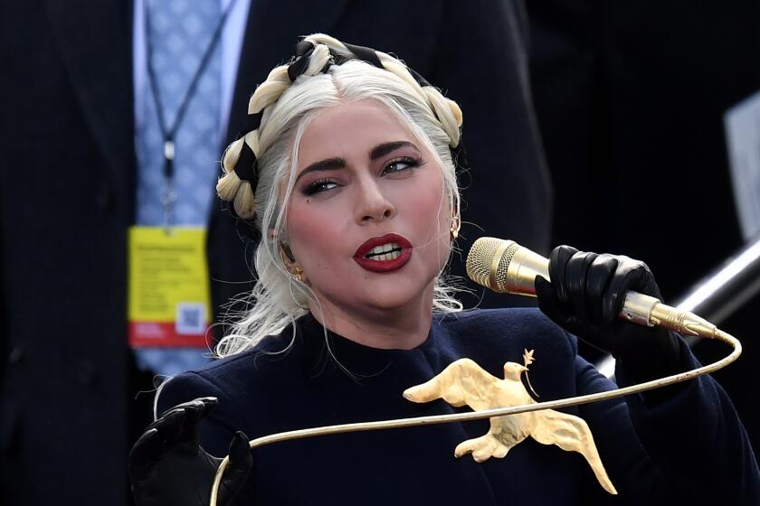 Lady Gaga singing into a golden microphone, wearing a black shirt and gold bird pendant