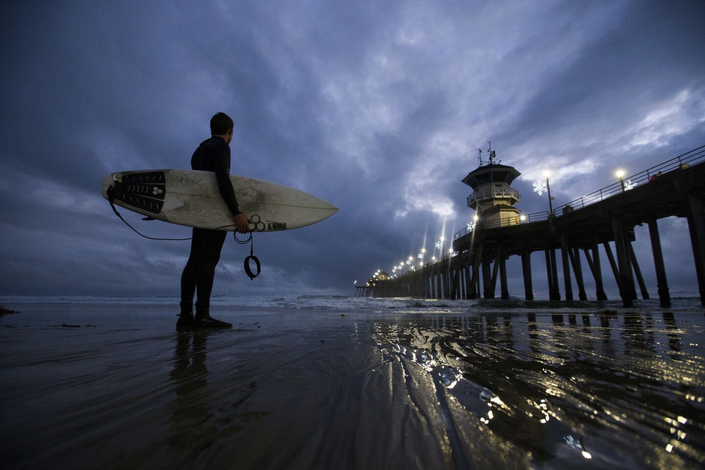 Surfer and Huntington Beach lifeguard Jachin Hamborg watches the dramatic sky and waves after surfing following his lifeguarding shift at dusk at the Huntington Beach pier.