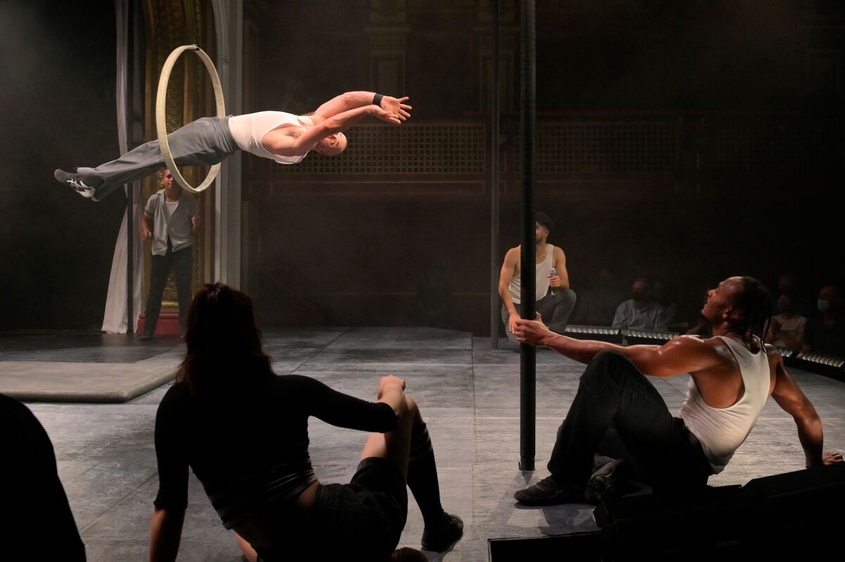 A man jumps headfirst through a suspended hoop as seated people watch.