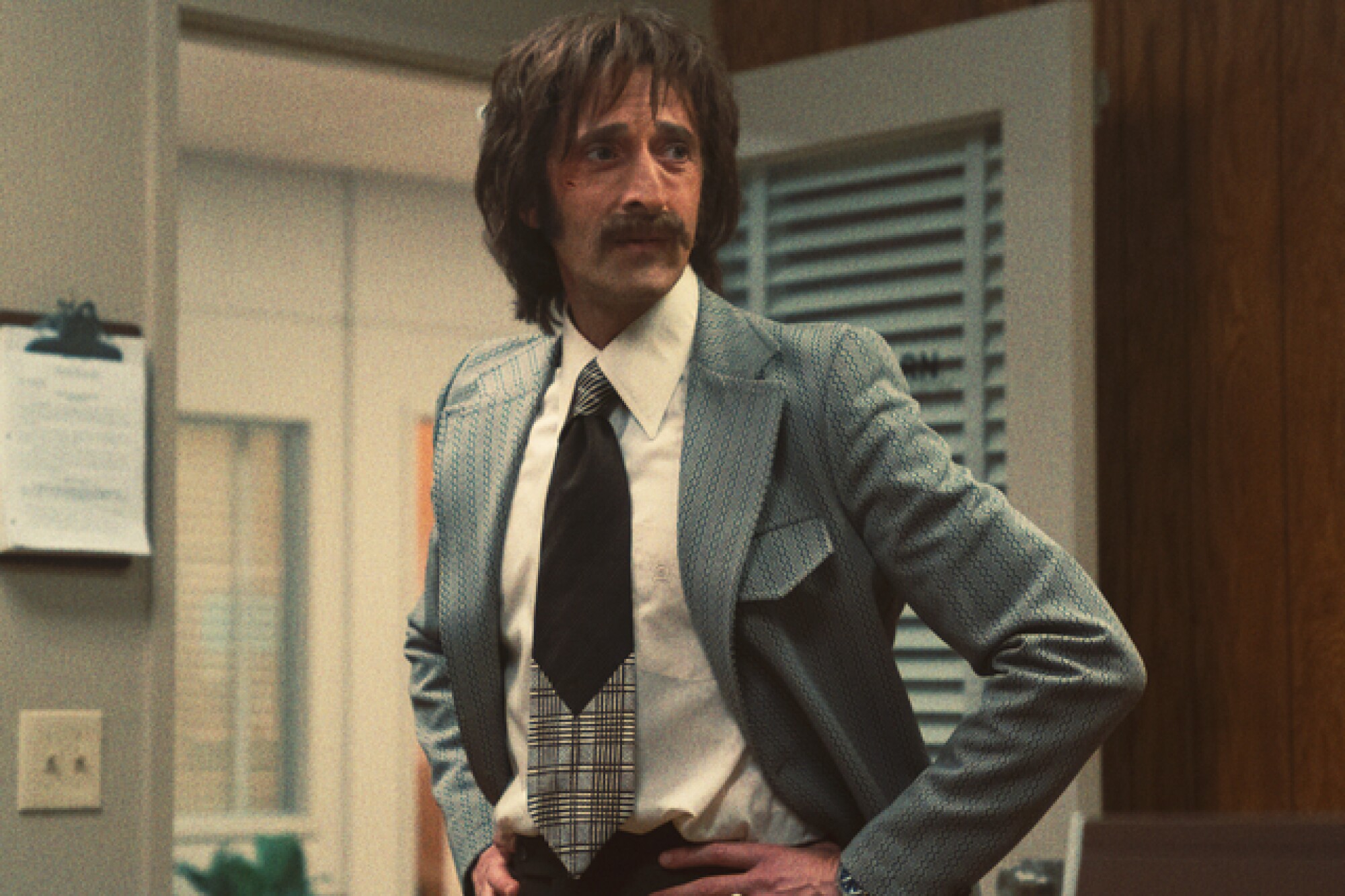 A man with shaggy hair and mustache wearing a suit with his hands on his waist