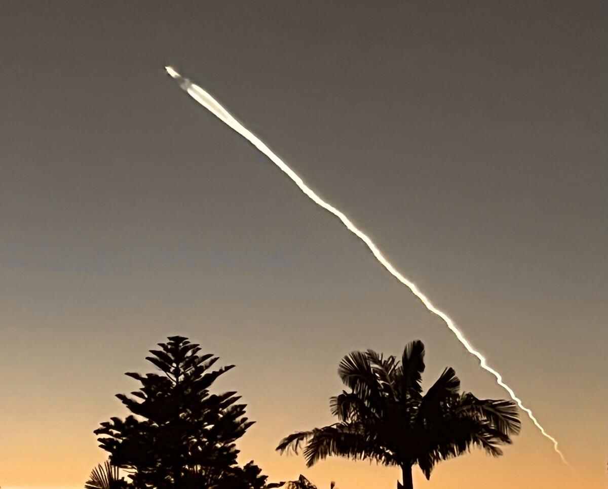 A rocket with a long contrail moves across a darkening sky above the outline of palm trees.
