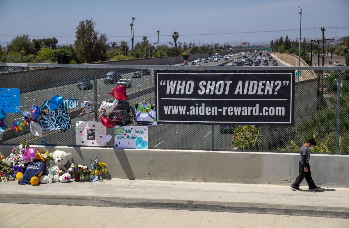A boy walks by a cluster of teddy bears, flowers and a sign that says "Who shot Aiden?" on a bridge overlooking the freeway.
