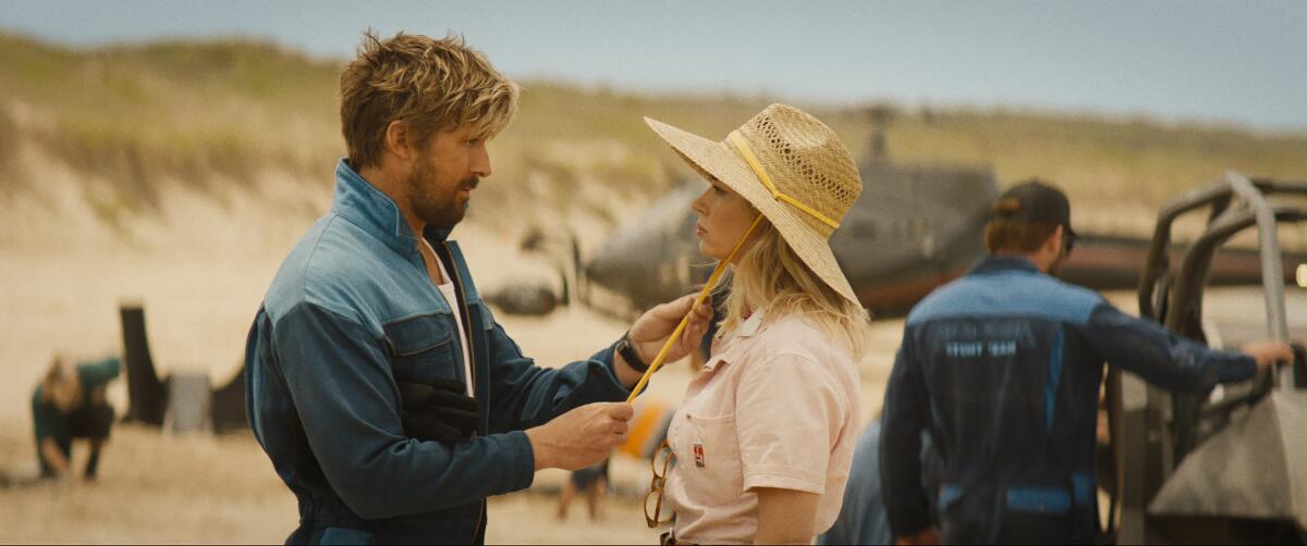 A man adjusts the strap of a woman's hat as they stand outdoors on a movie set's beach