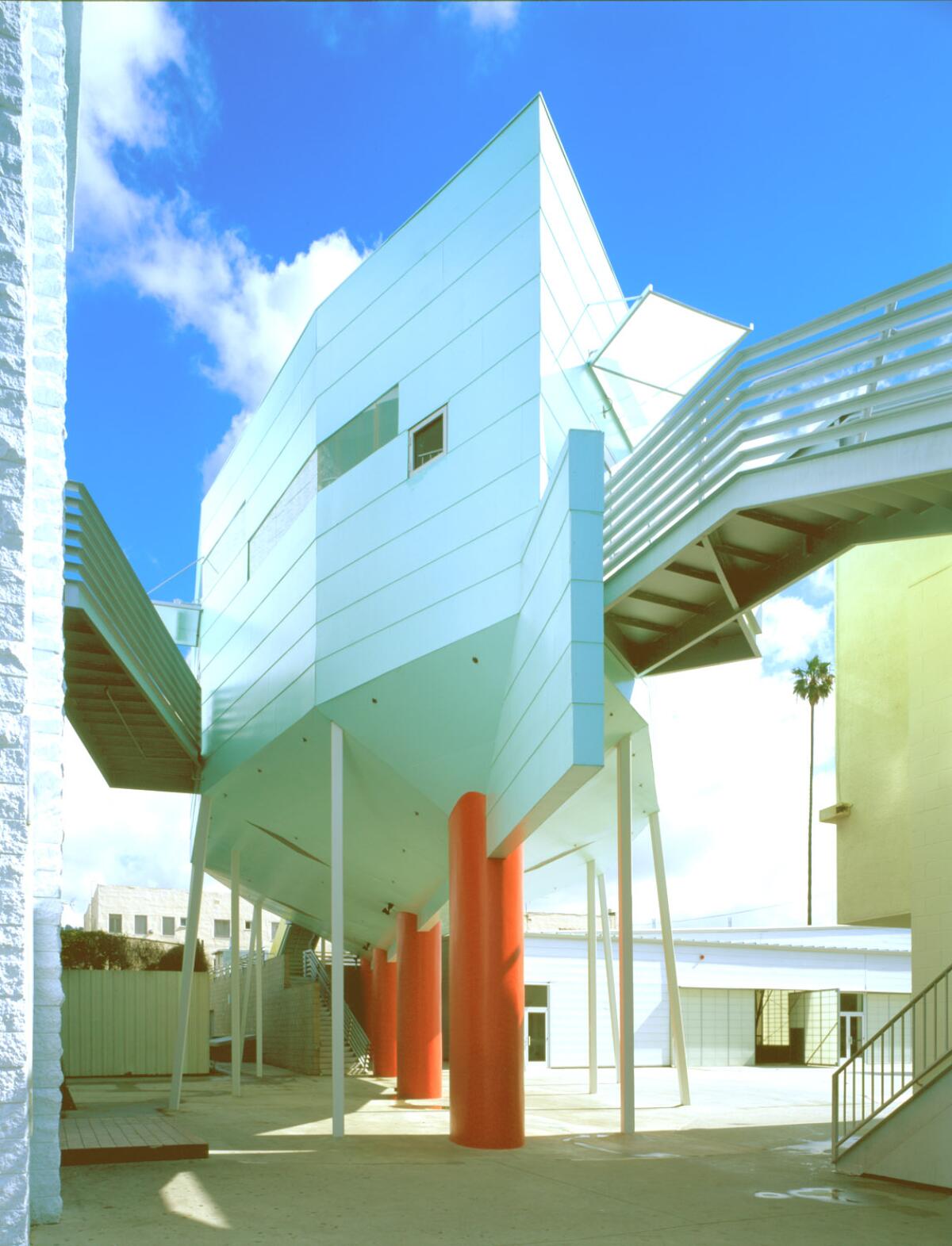 A light blue building whose form evokes a ship sits on red pilotis over a courtyard area