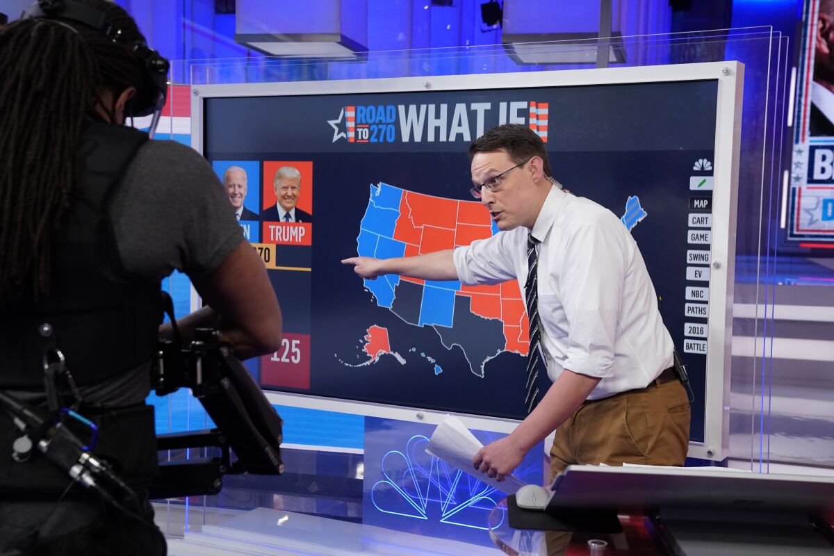 A cameraman films Steve Kornacki in front of a board with the words "What If," a U.S. map and images of Biden and Trump.