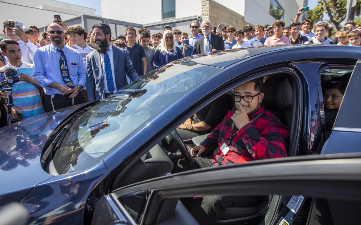 A teacher sits in a new car surrounded by students.