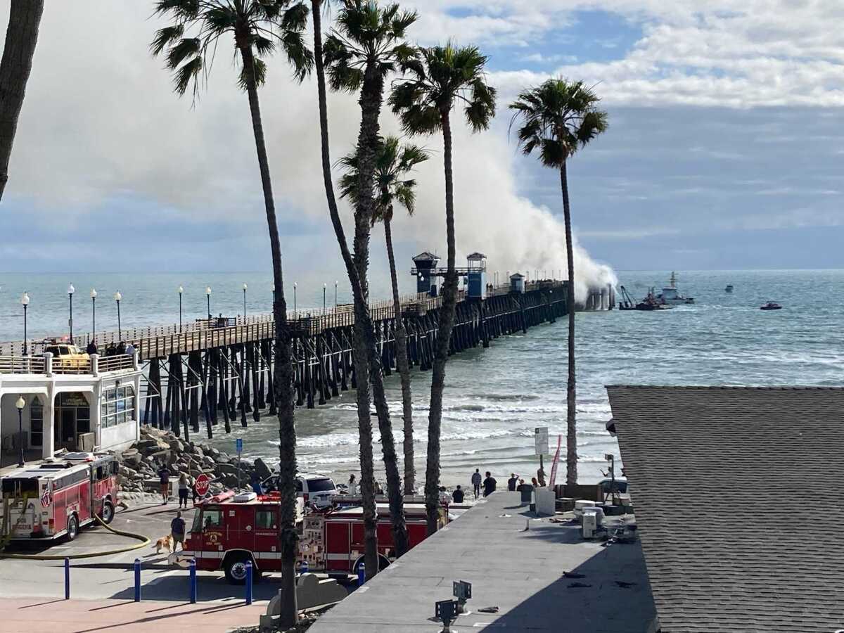 Smoke pours from the end of a pier as firetrucks are parked nearby