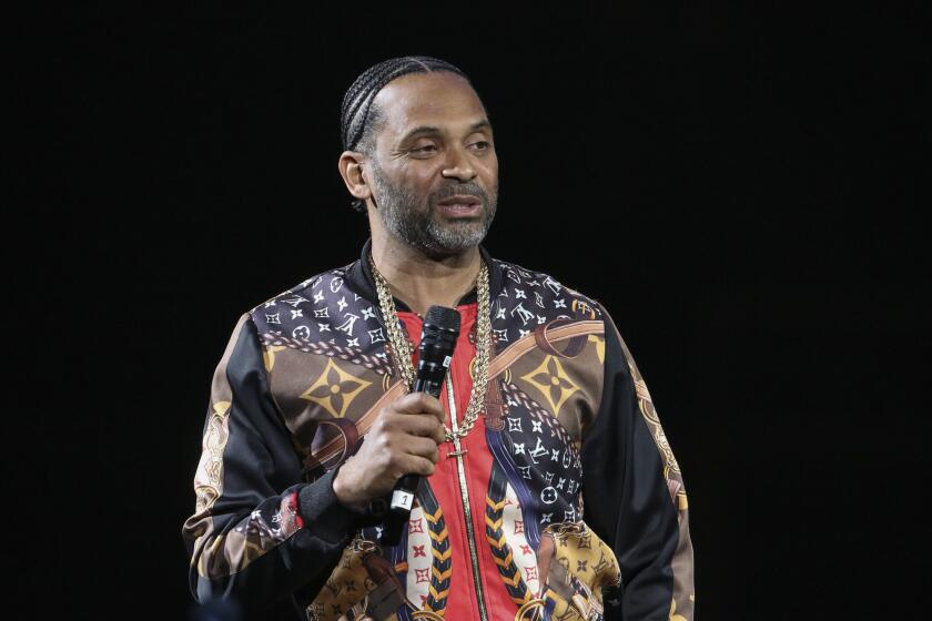 A man in a patterned jacket and gold necklace speaking into a microphone