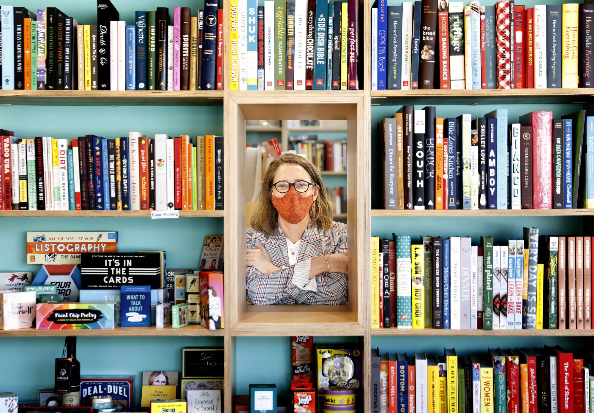 A woman in an orange mask peeking through a hole in a bookshelf.