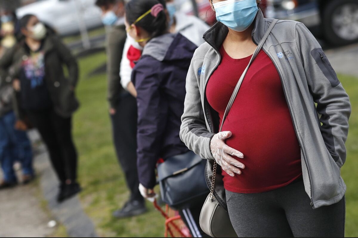 A pregnant woman wearing a face mask and gloves holds her belly as she waits in a line