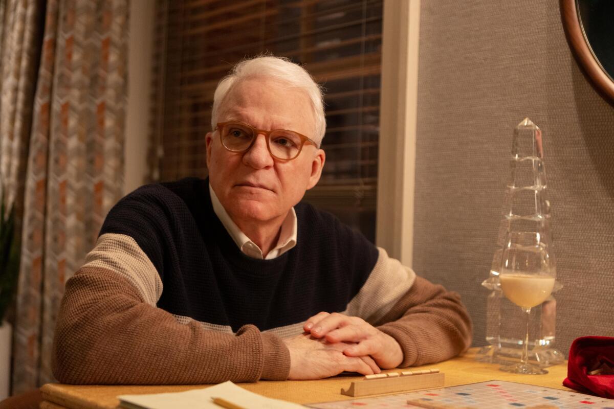 Steve Martin sitting at a table with his arms folded