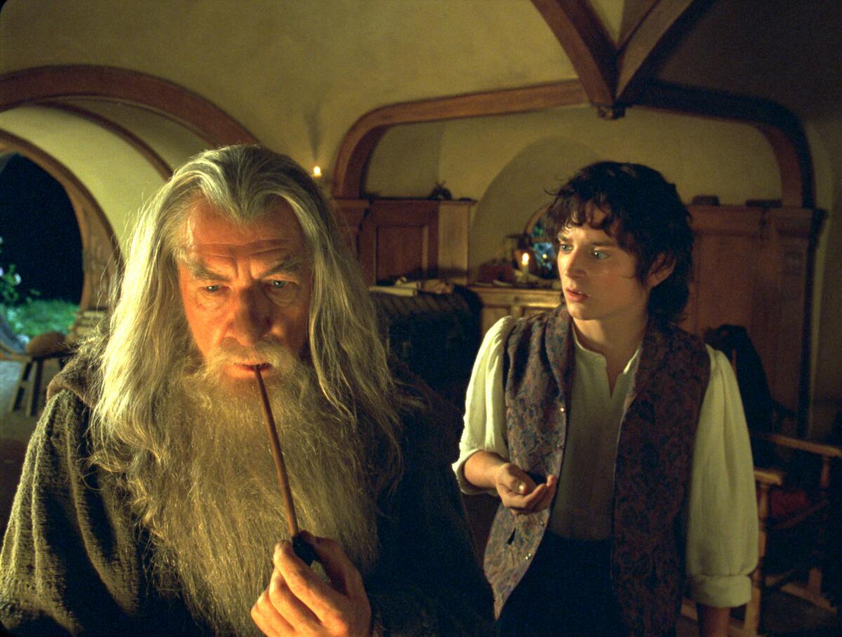A young hobbit stands behind a wizard with long gray hair and beard who's smoking a pipe