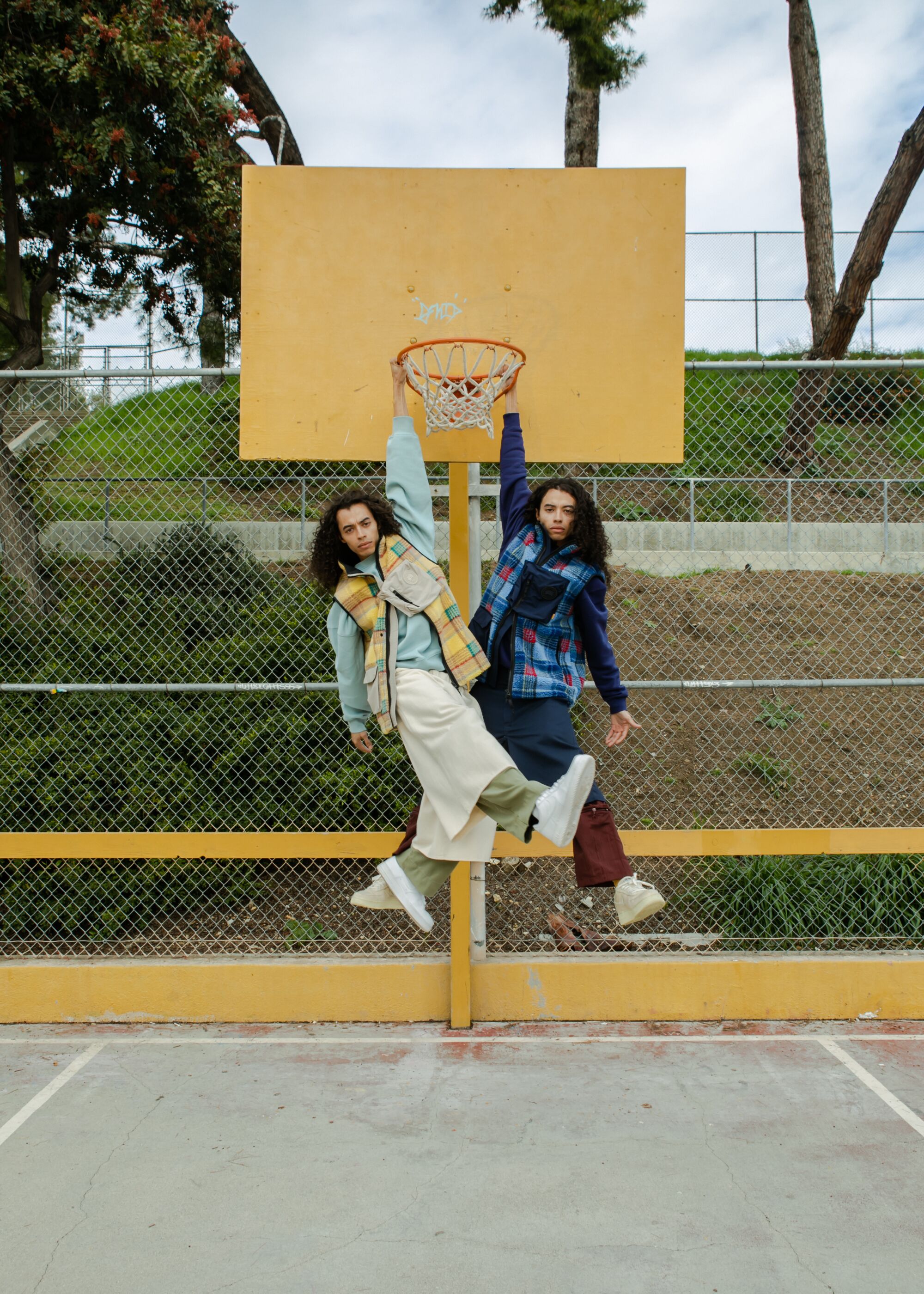 Two men hang from a basketball hoop while wearing tartan vests, one yellow, one blue.