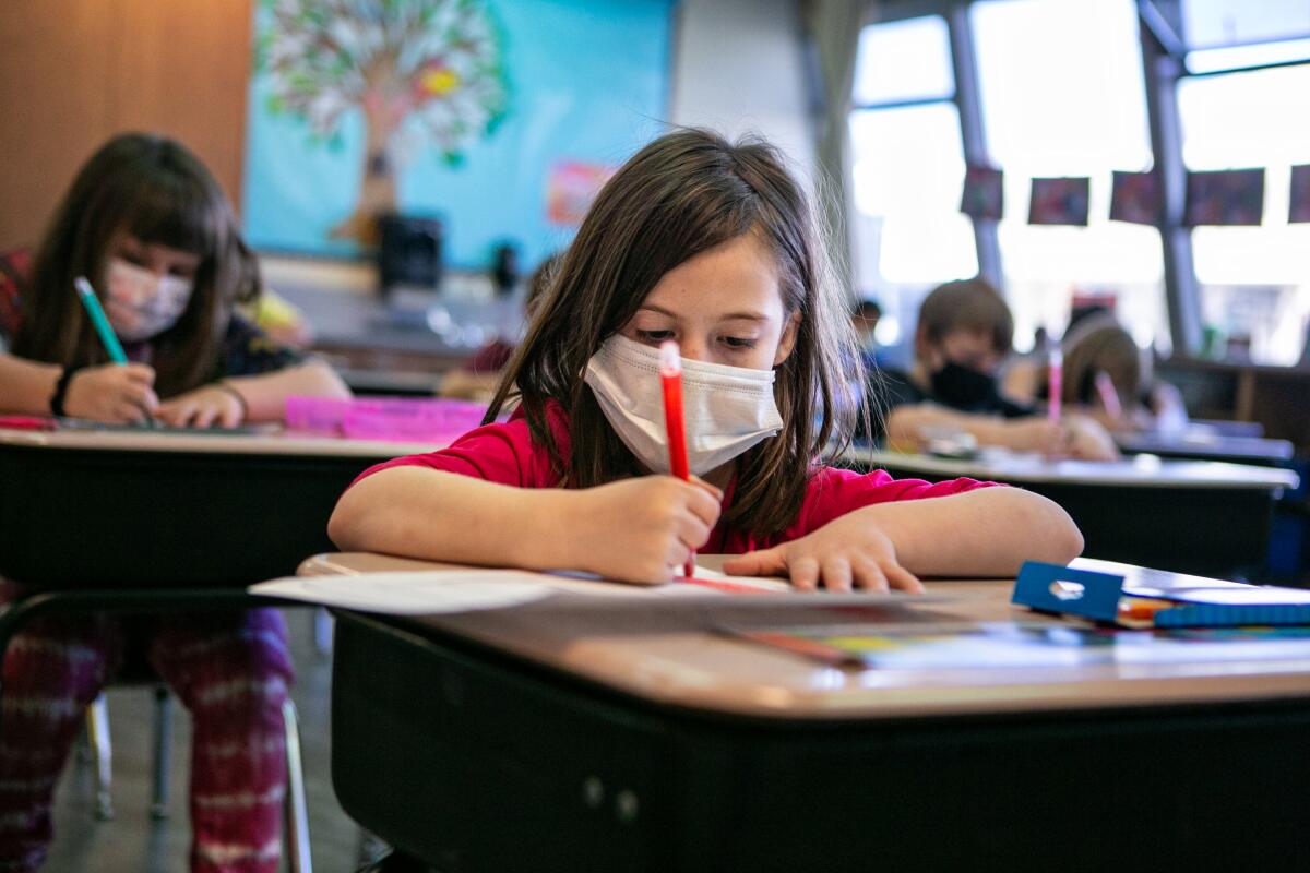 An elementary school girl wearing a white mask writes at her desk. 
