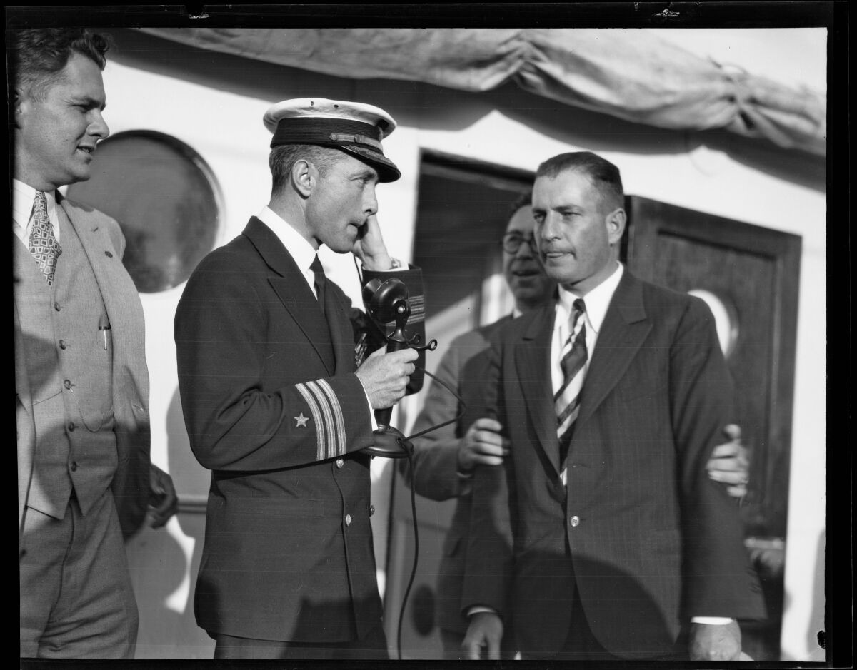 Several men, one in naval uniform, stand on the deck of a ship. One speaks into a candlestick phone.