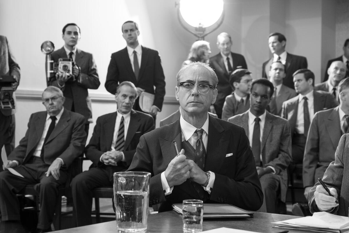 In a black-and-white scene, a man in a suit and horn-rimmed glasses sits at a table. Behind him are other men in suits. 