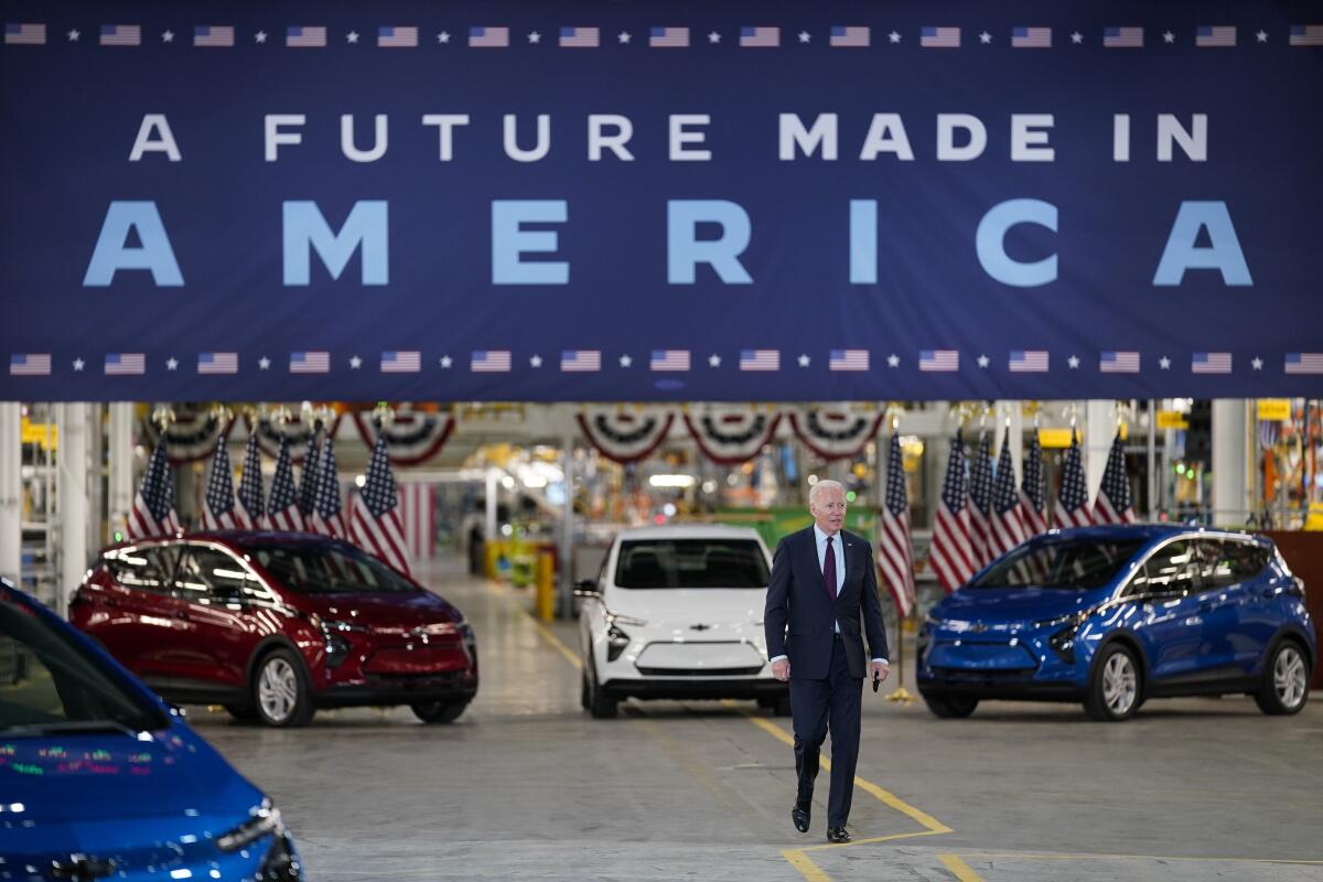 A man in a dark suit and tie walks in front of red, white and blue cars and a banner that says A Future Made in America 