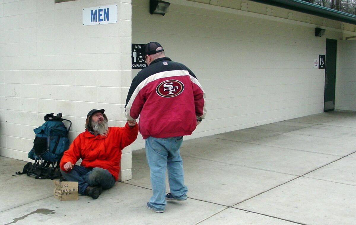 A man sitting with a backpack against a wall by a men's outdoor restroom, reaching up to receive something from another man