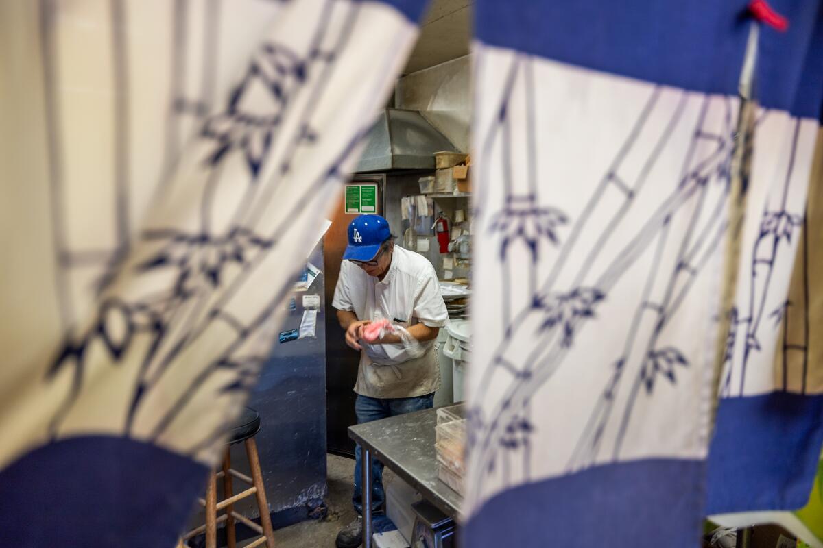 Mas Fujita holds pink mochi in his shop's kitchen, framed by blue-and-white fabric with illustrations of bamboo shoots