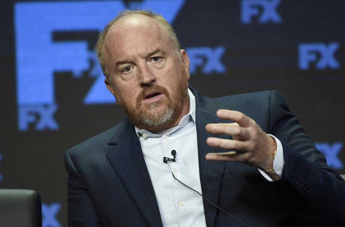 Louis C.K.in a black suit jacket, white suit shirt sitting and holding his left hand out