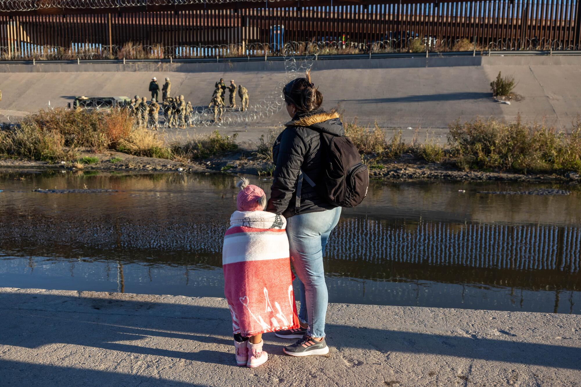 A woman and child look at uniformed men behind loops of concertina wire across a river channel 