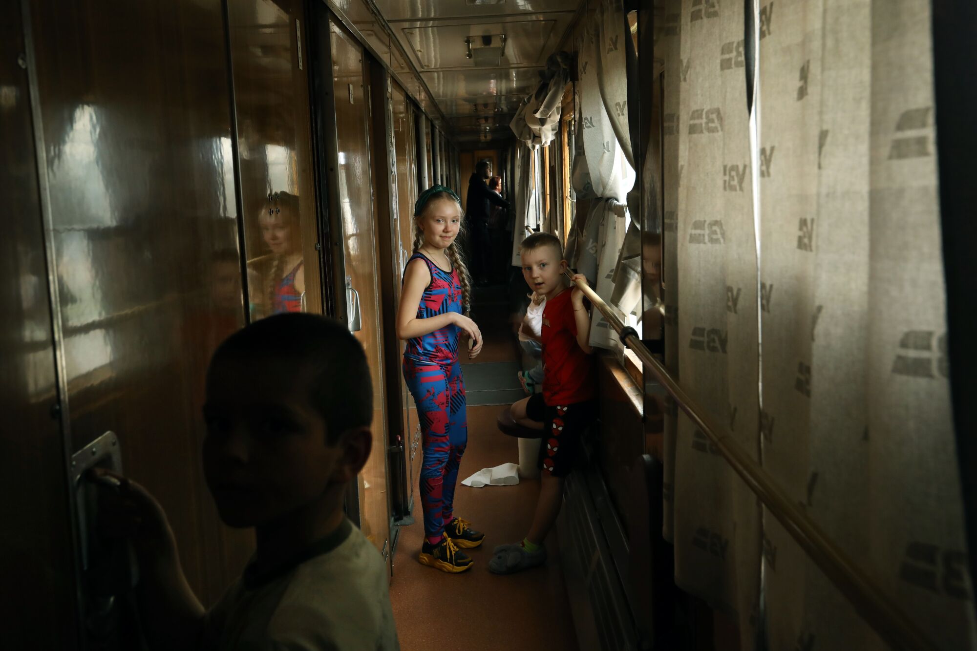 A girl and a boy stand in the train corridor while, in the foreground, a boy holds onto a door handle