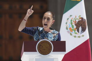 A woman in a dark top with blue flowers points with a finger as she speaks at a lectern near a green, white and red flag