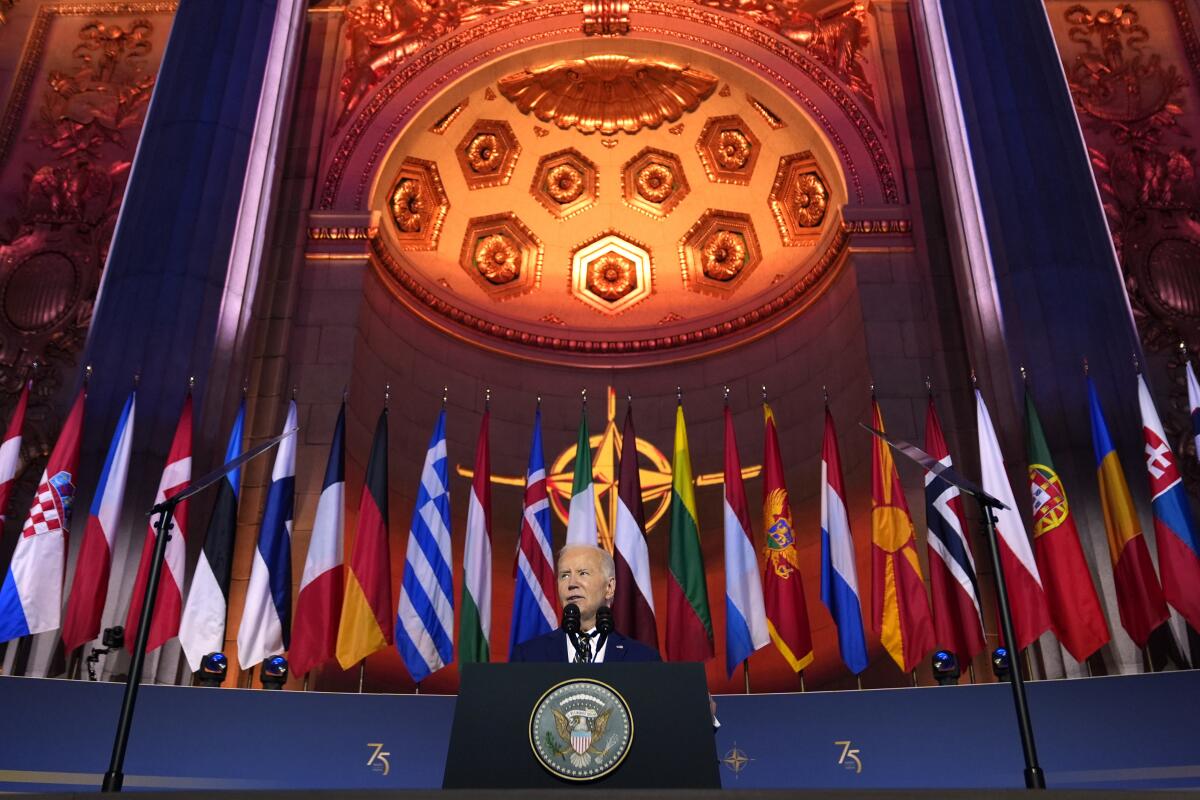 President Biden speaks from a lectern with the presidential logo below an ornate ceiling, a row of 2 dozen flags behind him