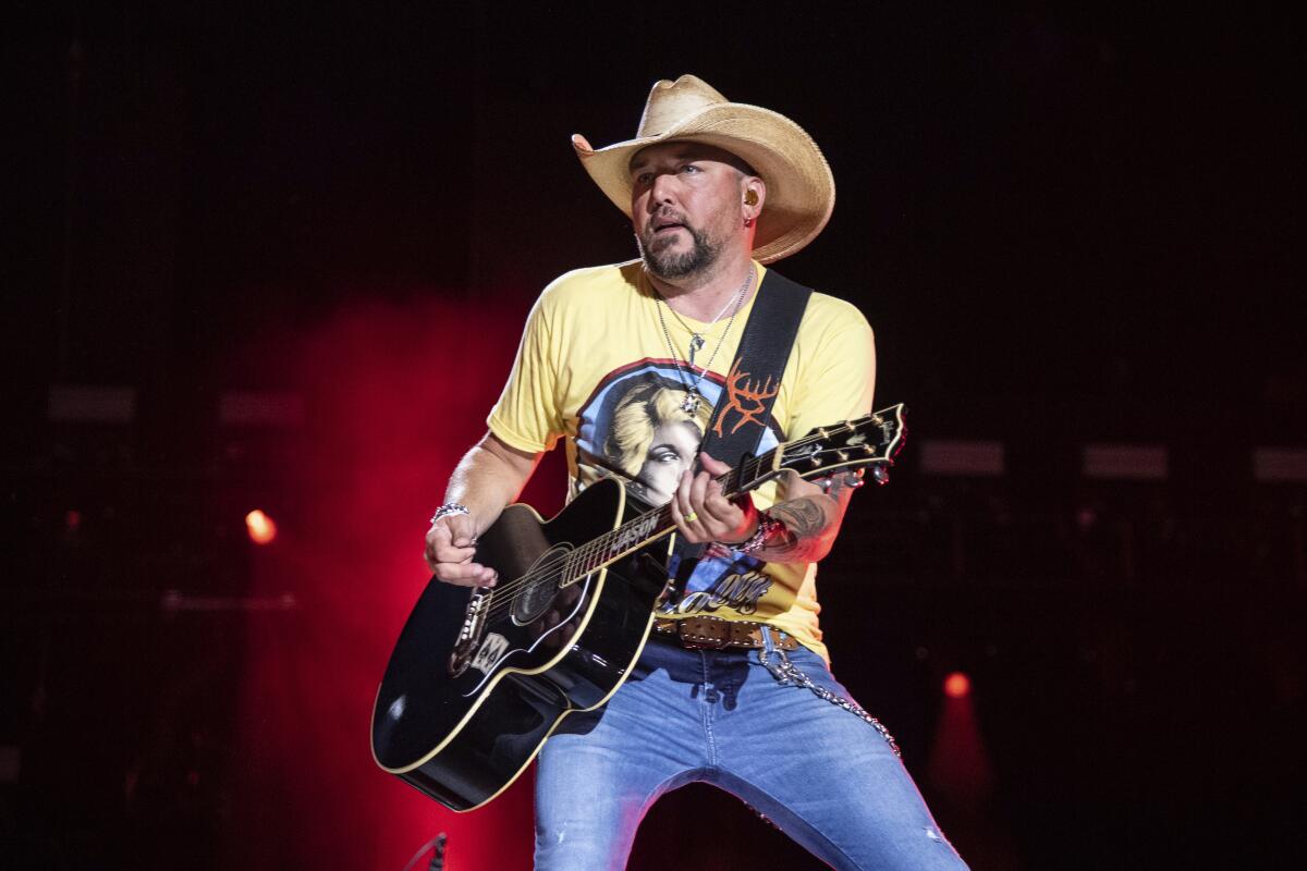 Jason Aldean wears a yellow shirt with a blue circle, blue jeans and a tan cowboy hat as he plays a guitar onstage