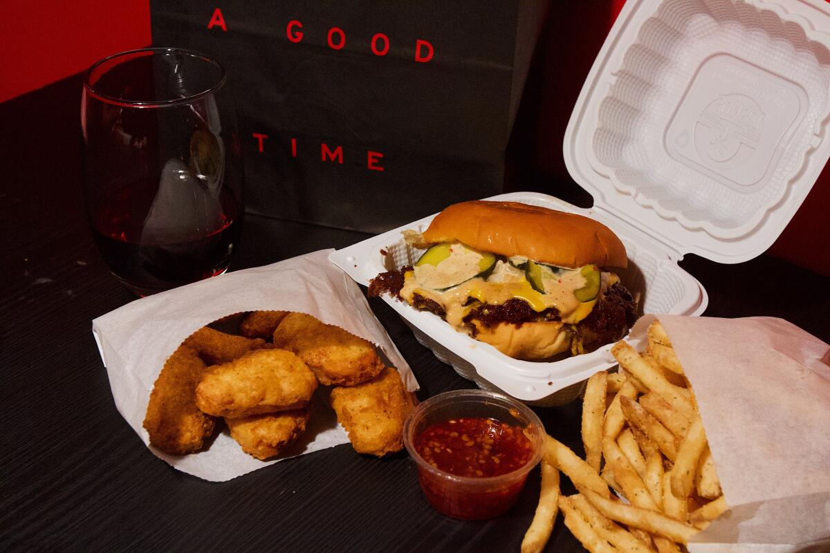 A cheeseburger, bag of chicken nuggets, bag of fries and glass of red wine on a black table