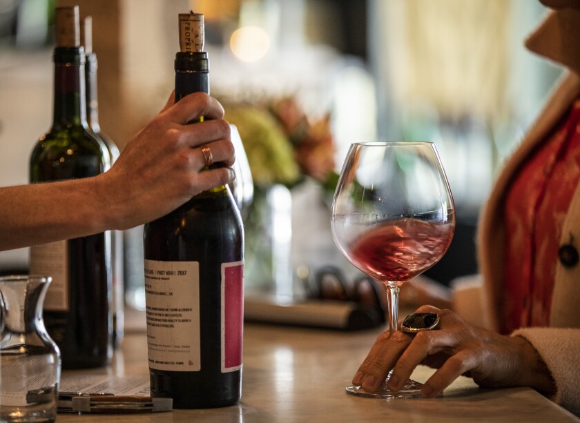 A hand holds the neck of a wine bottle and another hand holds the stem and base of a wine glass.