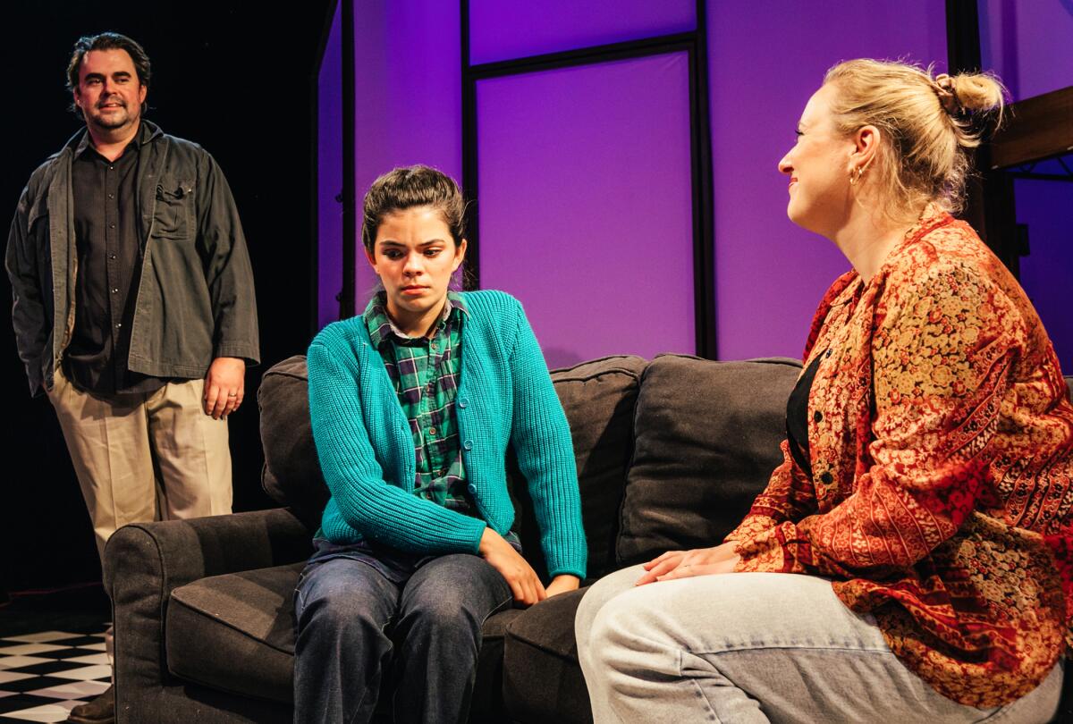 Two women sit on a sofa facing each other while a man in the background looks on