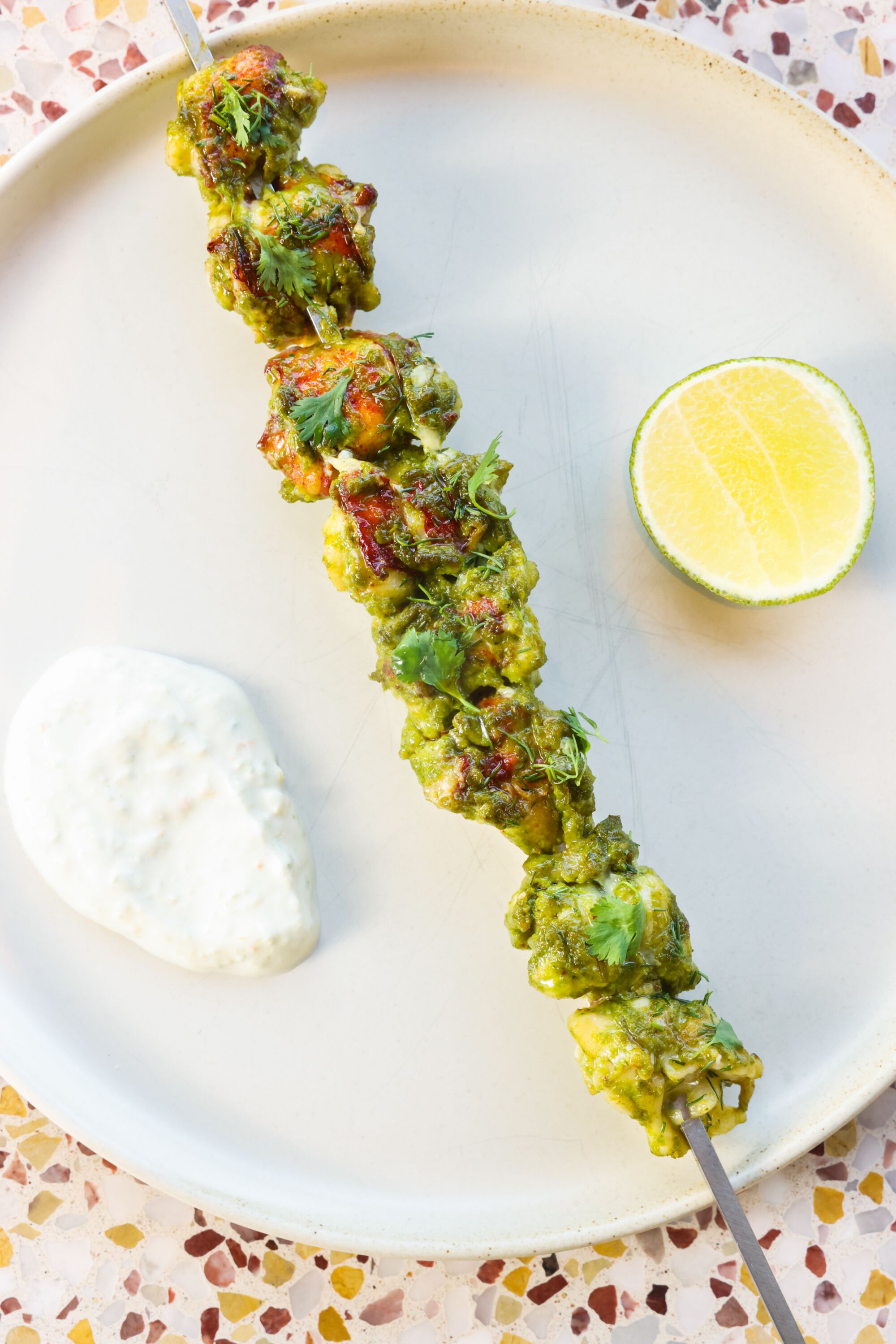 A grilled lobster skewer on a white plate, served with green harissa, serrano-lime yogurt and sprouting cilantro.