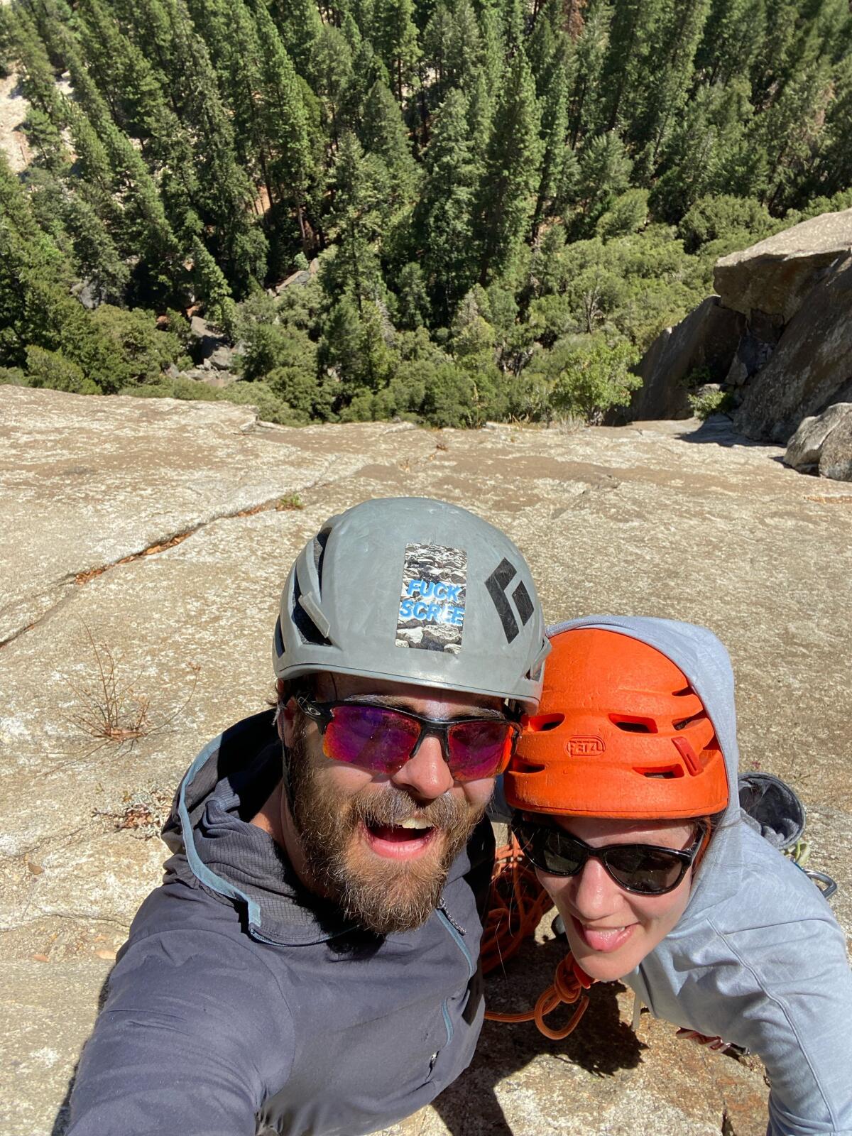 A man and a woman in climbing helmets smile for the camera from a cliff face with trees in the background. 