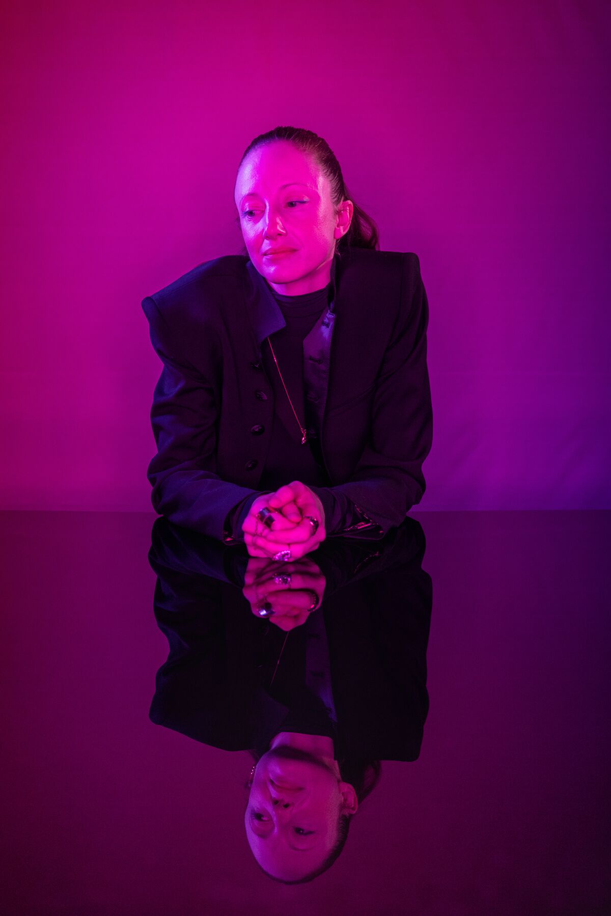 A woman sits at a table, her hands clasped in front of her, in a rosy magenta light.