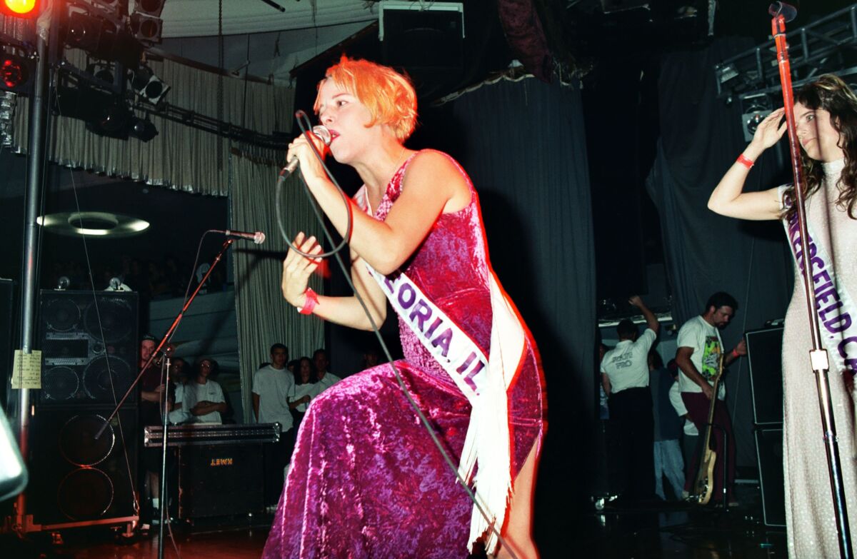 A female rock musician in a pink velvet dress and a sash performs onstage