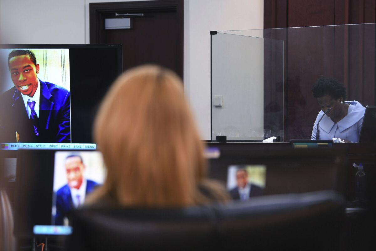 A woman in a courtroom while an image of a man is displayed nearby