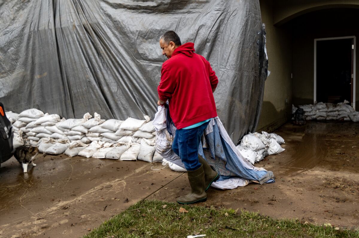 A man drags sheets down a wet driveway in front of a home covered by a tarp with stacks of sandbags outside.