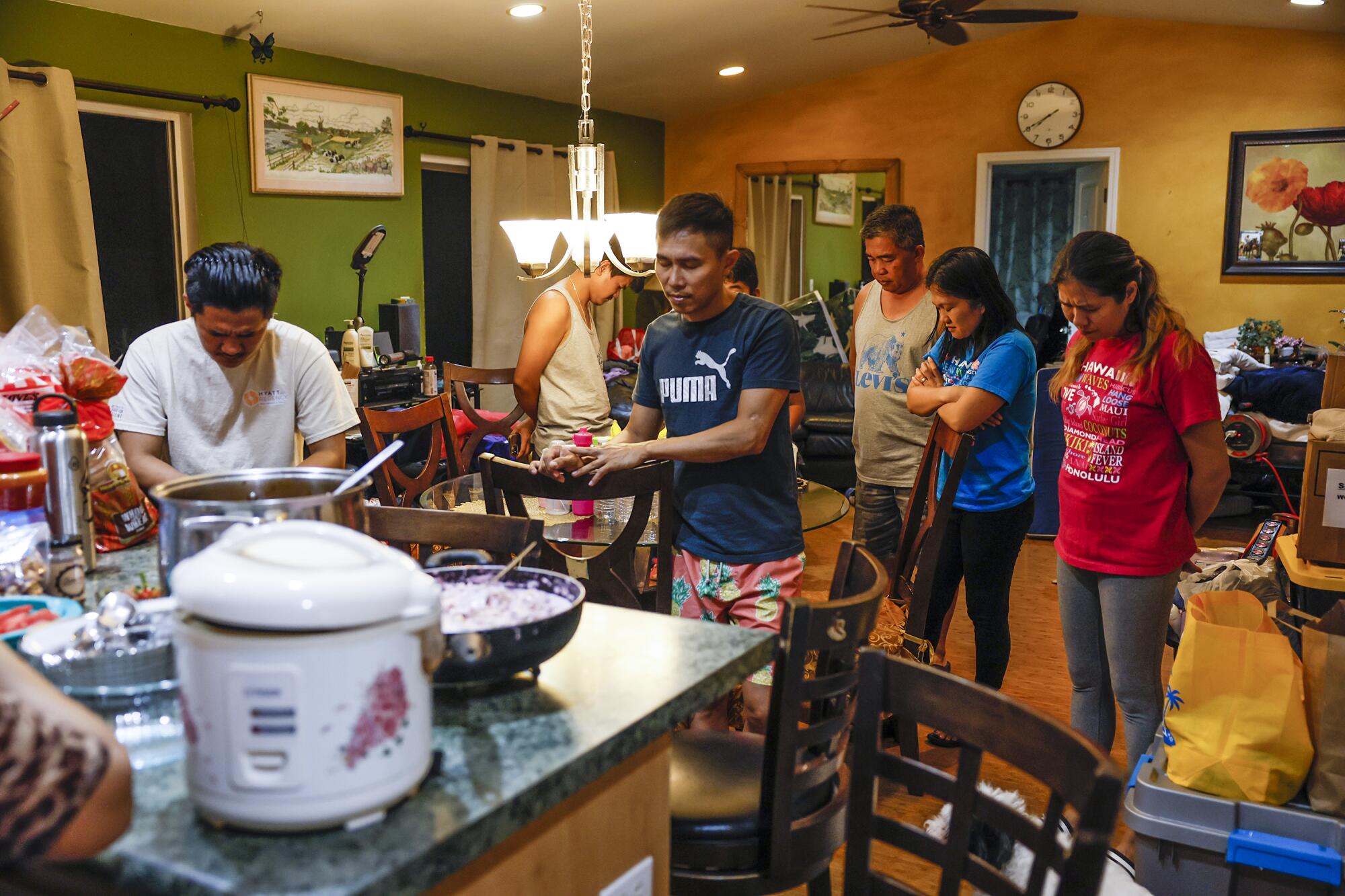 People stand with heads bowed before a kitchen counter, where a rice cooker and pots and pans sit