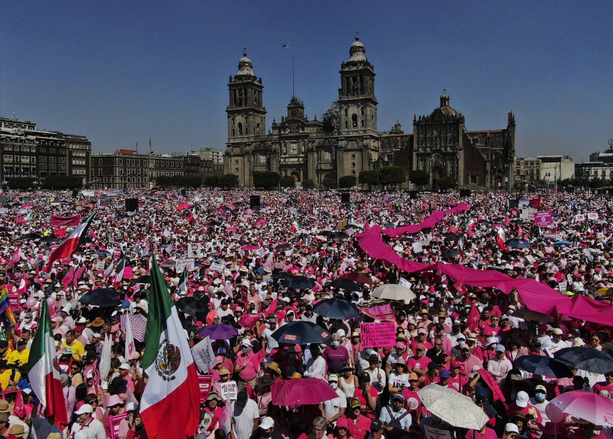 Binaların arka planında Meksika'nın yeşil, beyaz ve kırmızı bayrağını ve pembe pankartlarını sallayan büyük kalabalıklar görülüyor.