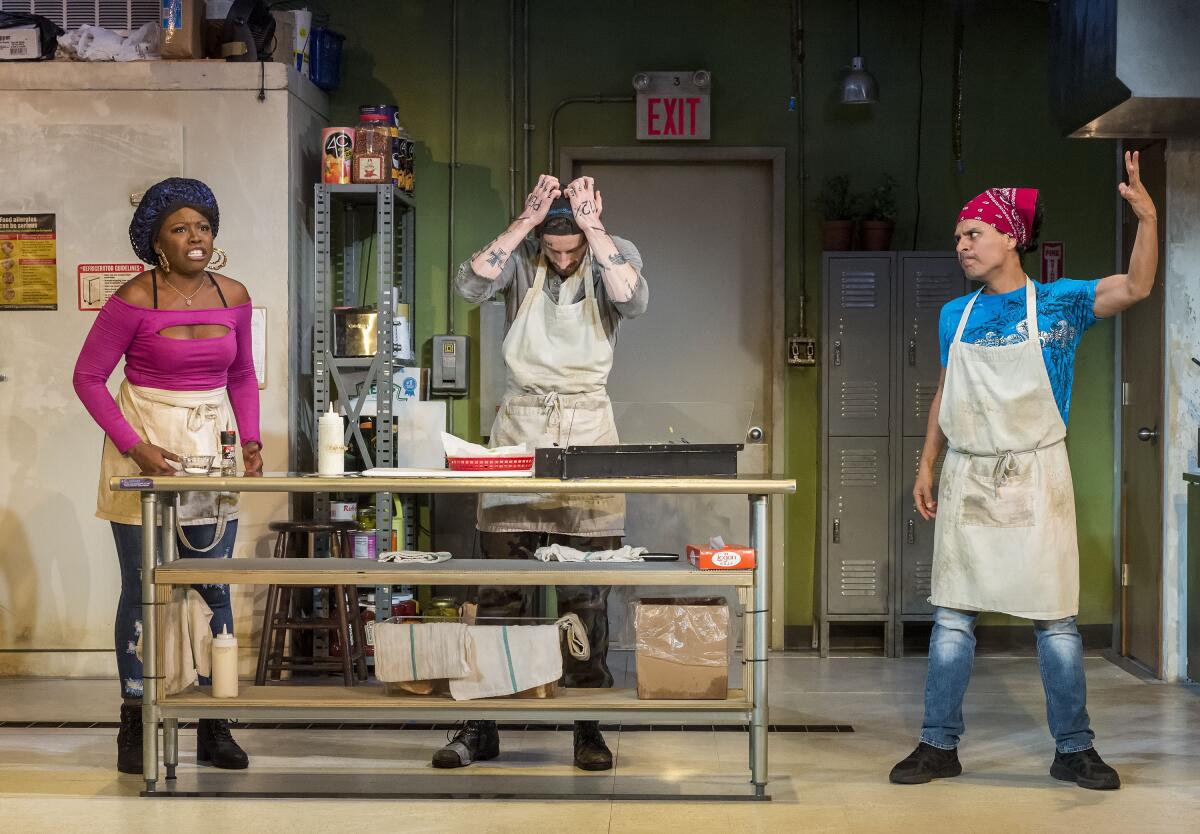 Three workers in a sandwich shop kitchen talk animatedly in a scene from "Clyde's" at the Mark Taper Forum.