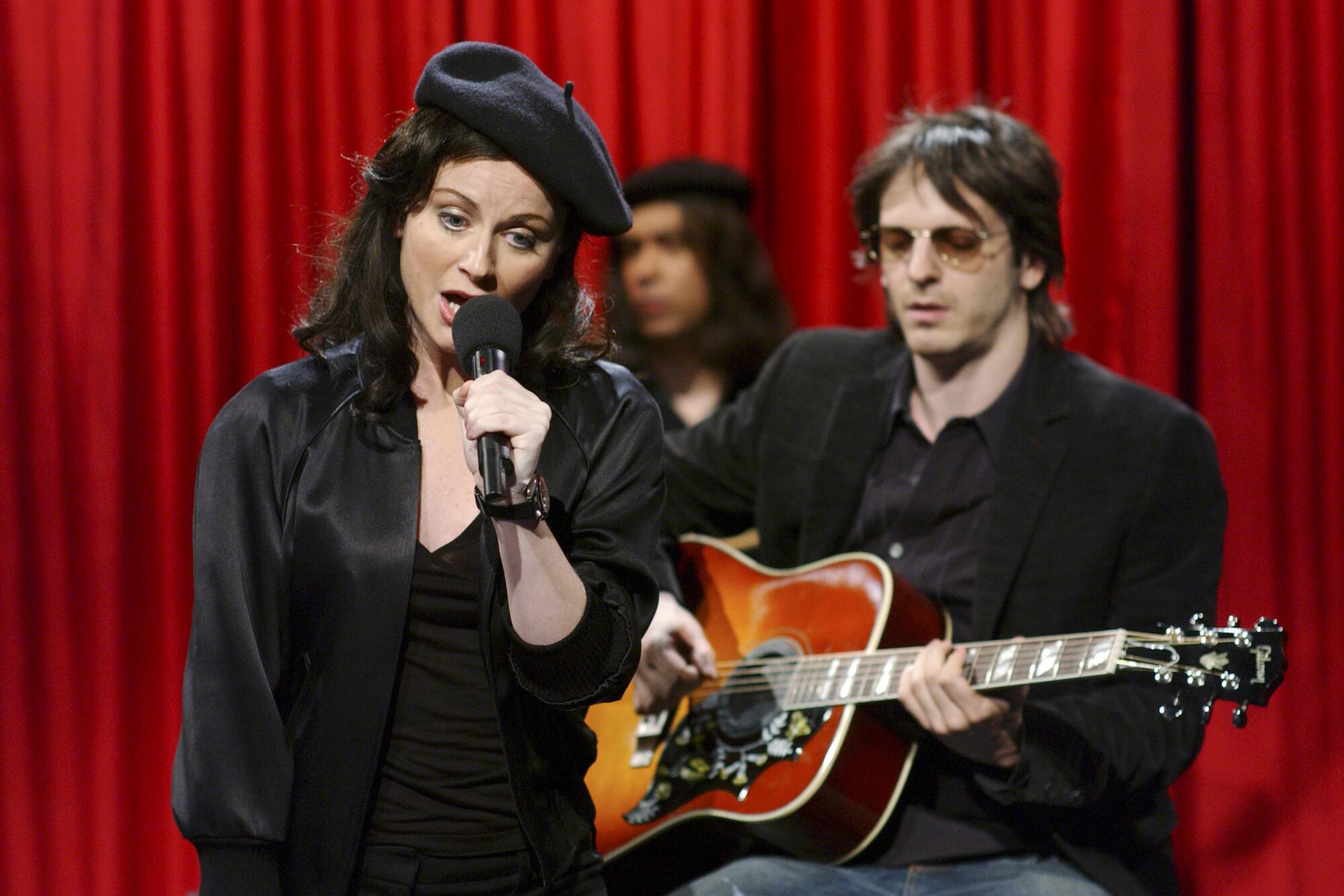 A singer (Amy Poehler as Madonna) dressed in black with a black beret in front of a guitarist (Dr. Luke) wearing sunglasses.