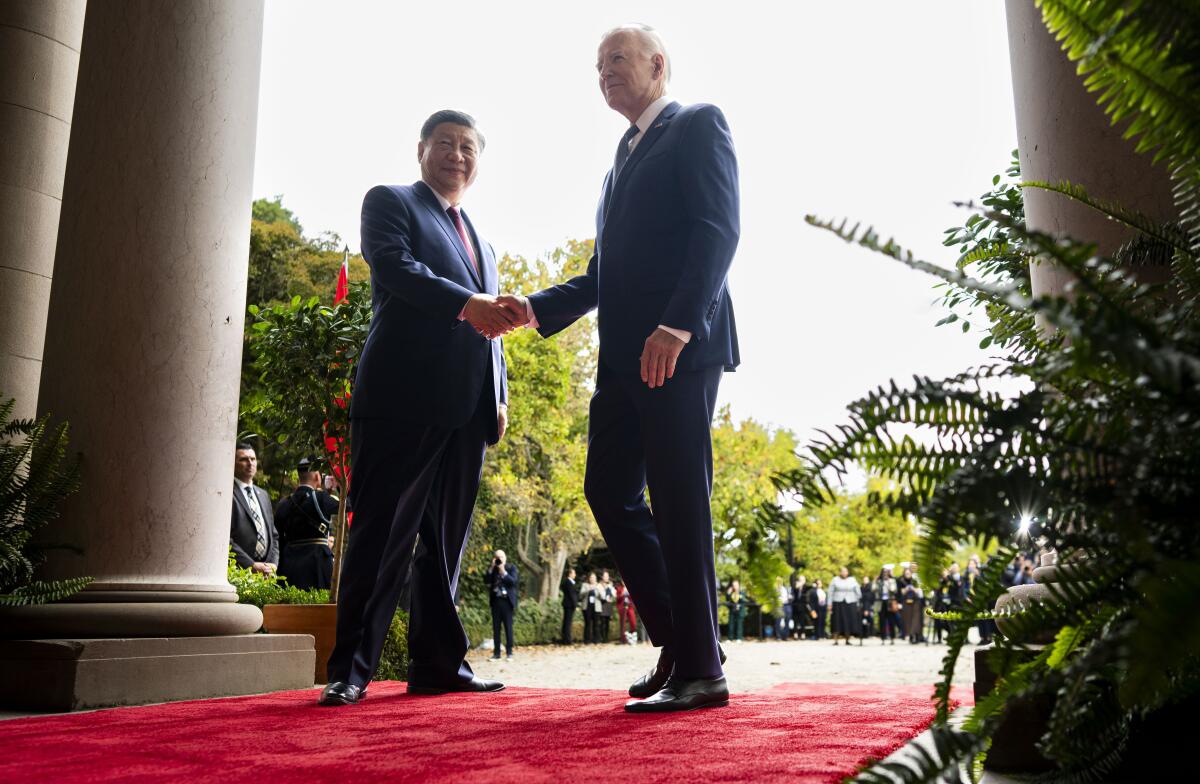 Two men in dark suits and ties shake hands while standing on a red carpet 