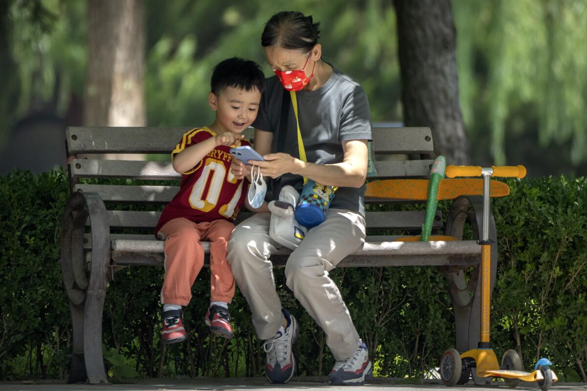Una mujer con mascarilla y un niño observan un celular sentados en una banca en un parque público