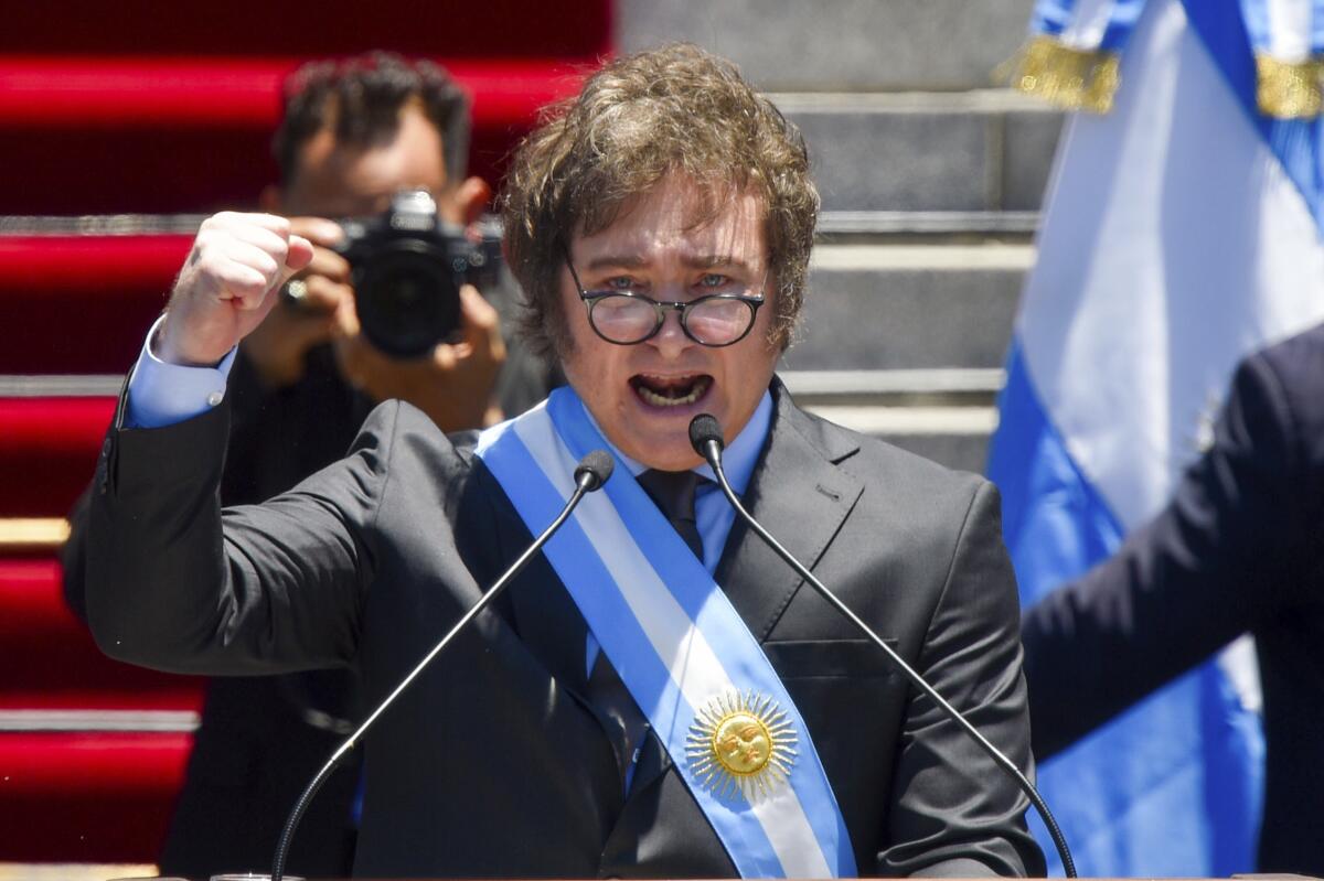 President Javier Milei holds up a fist and speaks outside while wearing a blue and white sash with a sun image on it.