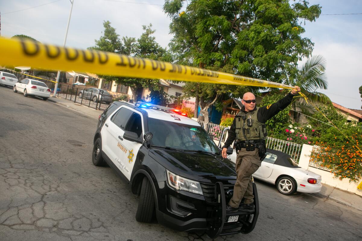 A police officer holds up some yellow police tape while standing on the bumper of a police vehicle in a residential street