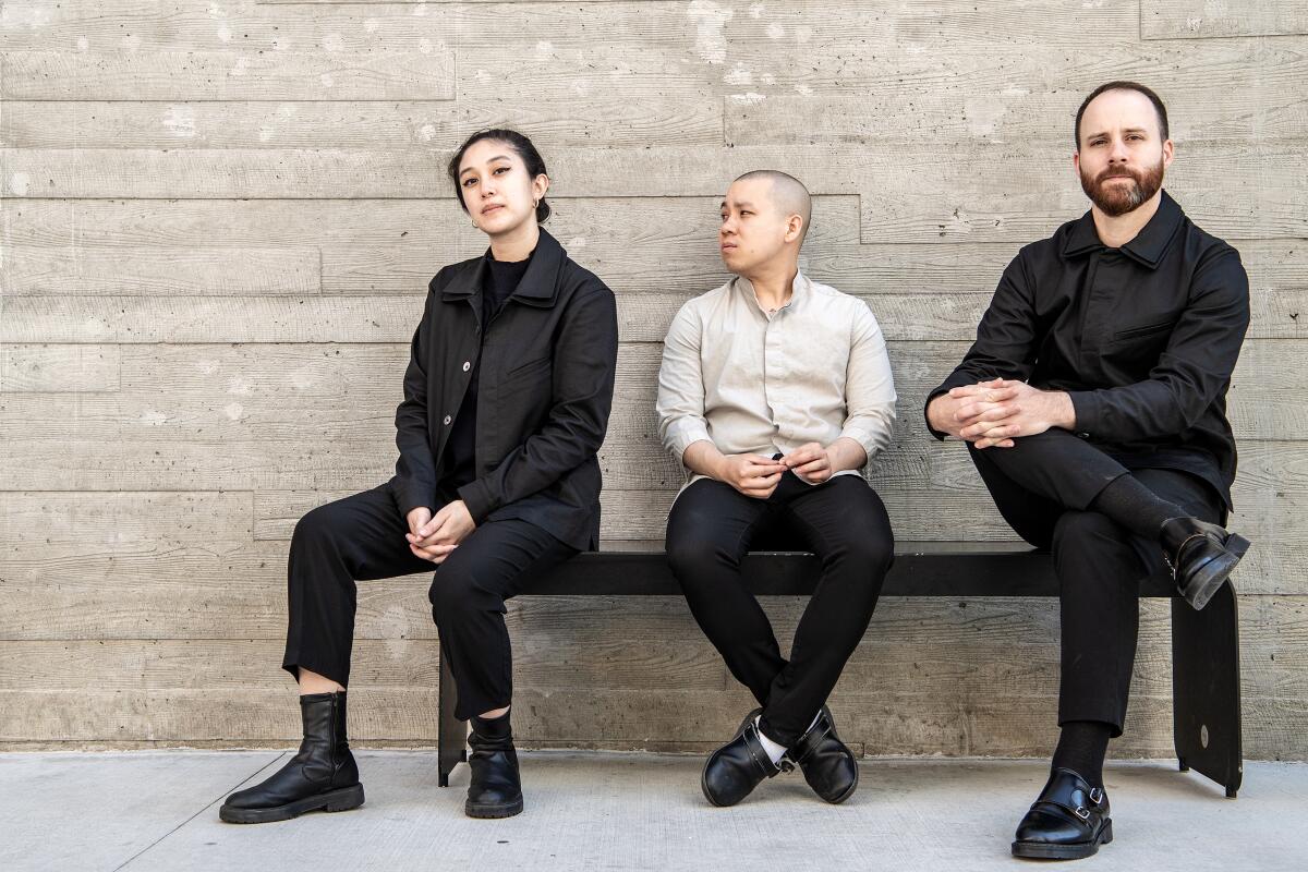 Three people sit on a bench in front of a gray wall.
