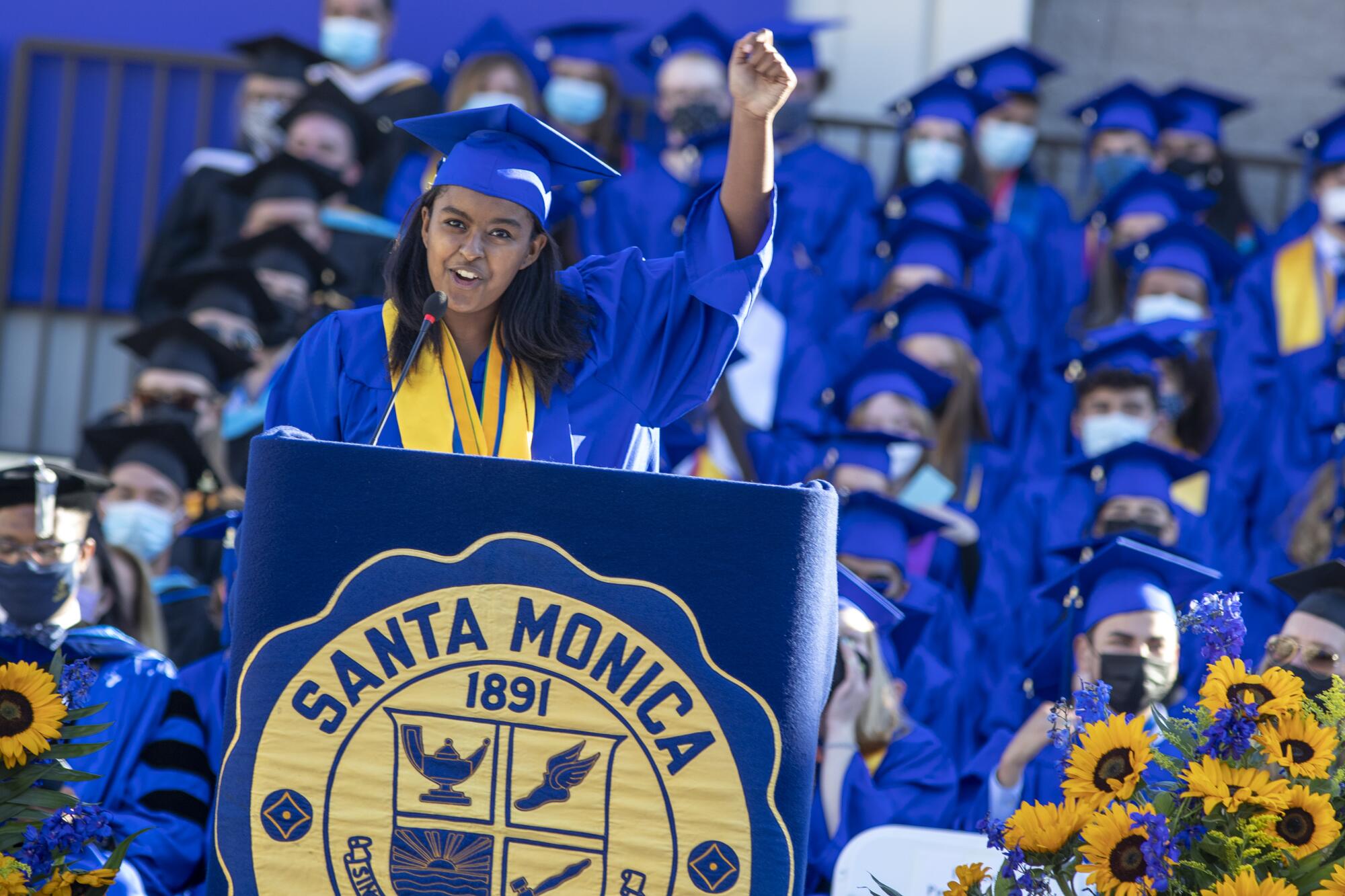 A high school graduate in cap and gown speaks into a microphone at a lectern and lifts one fist in the air.