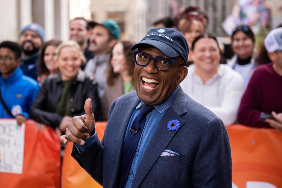 Al Roker smiles and gives a thumbs up in front of a barricaded crowd. He wears a blue suit and ball cap.